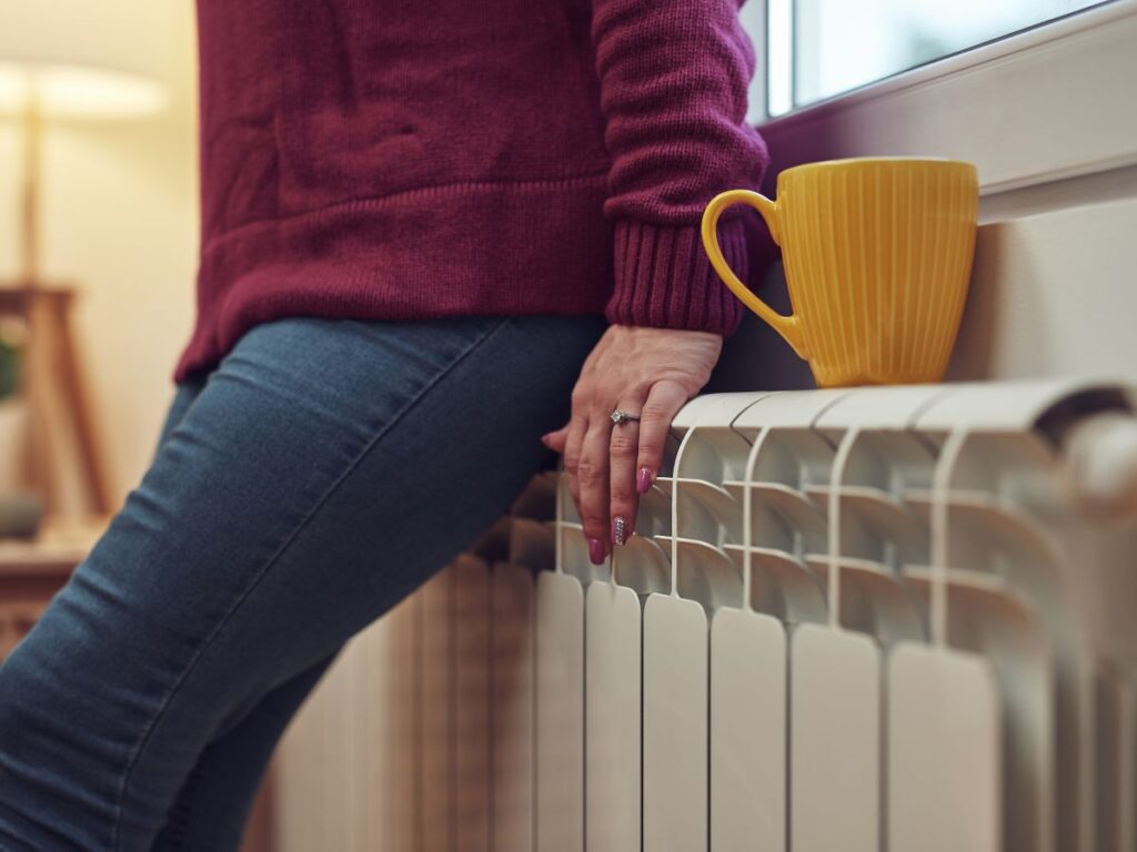 A person staying warm, leaning against a heater in a cozy home. This image illustrates the types of Medford heating services provided by Lund Plumbing and Heating to homes and businesses in Medford, MA