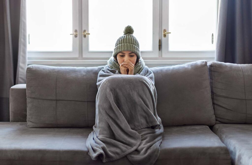 An illustration the need heating services in Tewksbury. A woman bundled up on couch because her heat isn't working,