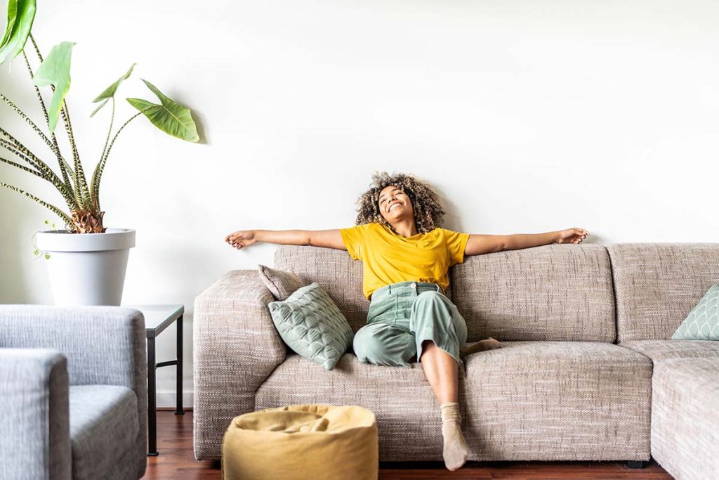 Cheerful woman stretching on a sofa with her arms wide open in a well-lit living room, highlighting Lund Plumbing and Heating's cooling services in Andover, Massachusetts.