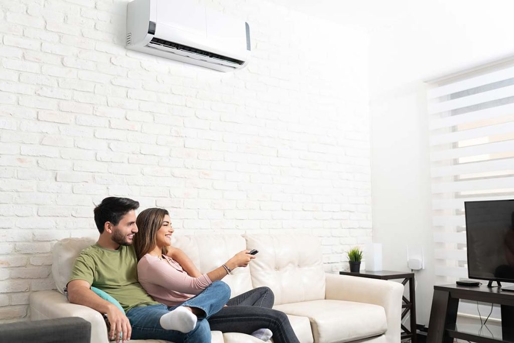 A happy couple relaxing on a couch under a wall-mounted air conditioner, enjoying the comfortable climate provided by Lund Plumbing and Heating's cooling solutions in Malden, Massachusetts.