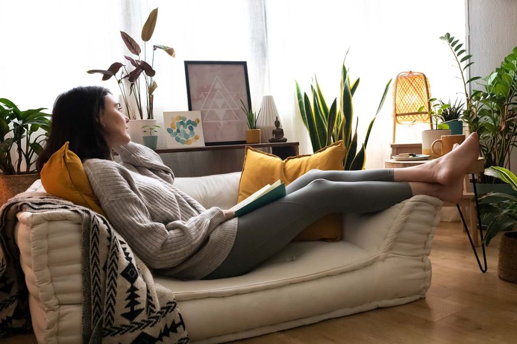 Relaxed woman lounging on a daybed with a book, surrounded by lush indoor plants, embodying customer comfort and satisfaction with Lund Plumbing and Heating in Andover, Massachusetts.