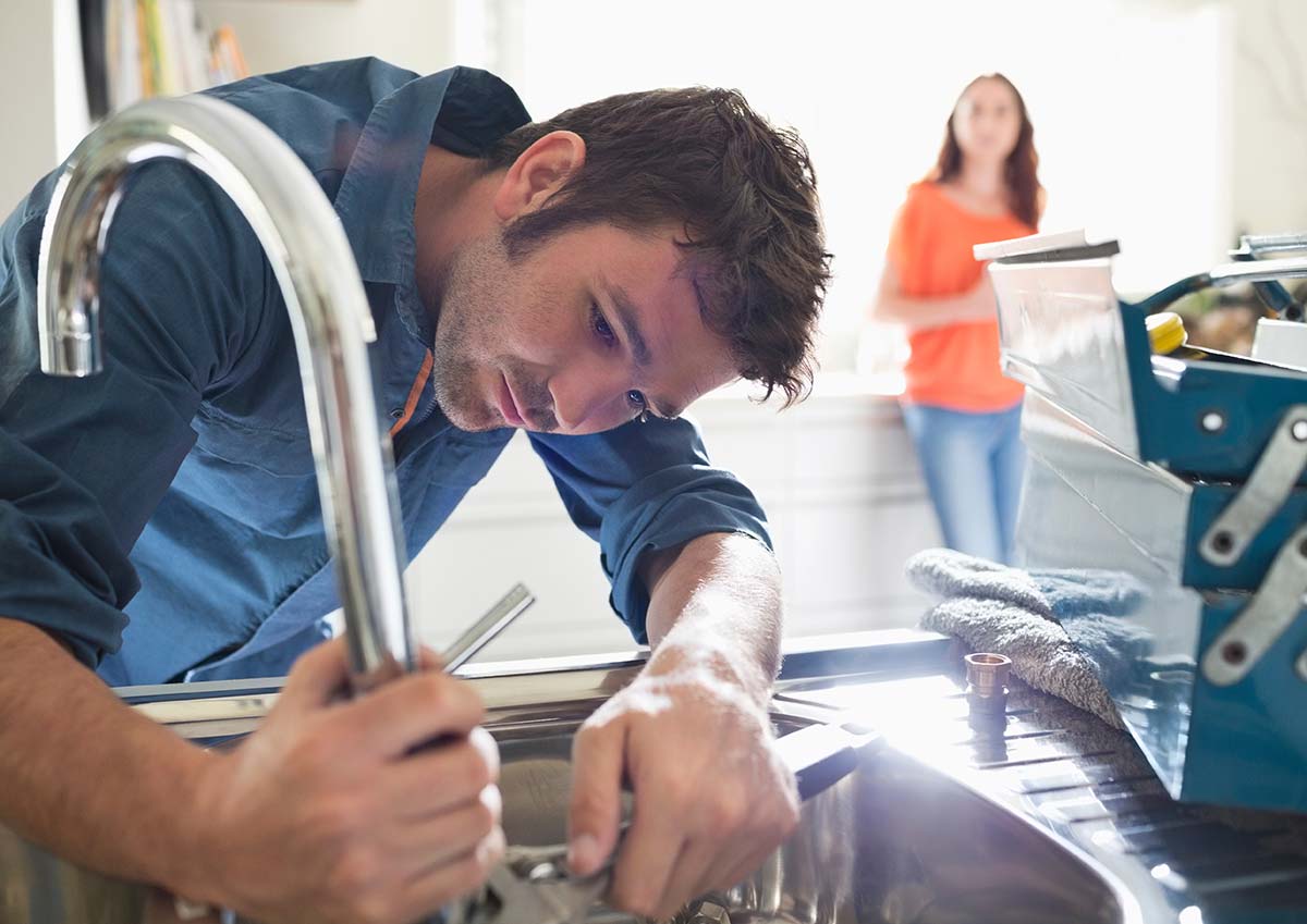 A kitchen plumber fixing a faucet with a wrench, with a customer watching in the background.